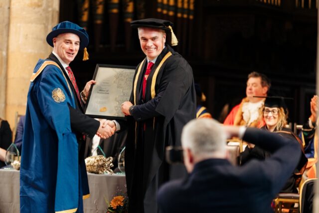 Two men in academic regalia smiling and shaking hands at a graduation ceremony. One holds a diploma. An audience applauds in the background.