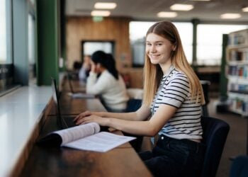 A young woman in a striped shirt smiles while using a laptop in a library. Open books and shelves are visible, conveying a study-focused atmosphere.