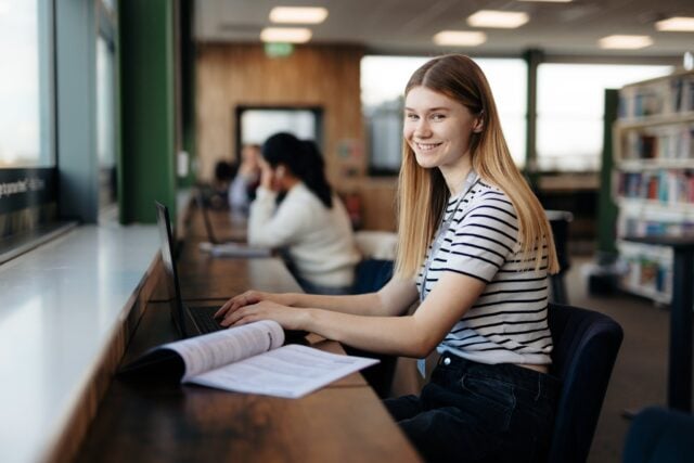 A young woman in a striped shirt smiles while using a laptop in a library. Open books and shelves are visible, conveying a study-focused atmosphere.