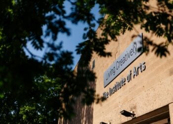 Sign reading "Living Creatively, The Institute of Arts" on a tan building, partially obscured by leafy green trees, with a clear blue sky above.