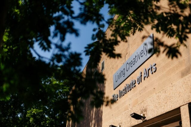 Sign reading "Living Creatively, The Institute of Arts" on a tan building, partially obscured by leafy green trees, with a clear blue sky above.