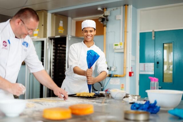 Two chefs in a kitchen; one smiling while piping dough with a blue bag, the other focused on kneading dough. Atmosphere is cheerful and busy.