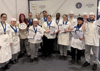 A group of smiling chefs in white uniforms and striped pants proudly display medals and certificates against a backdrop with event logos.