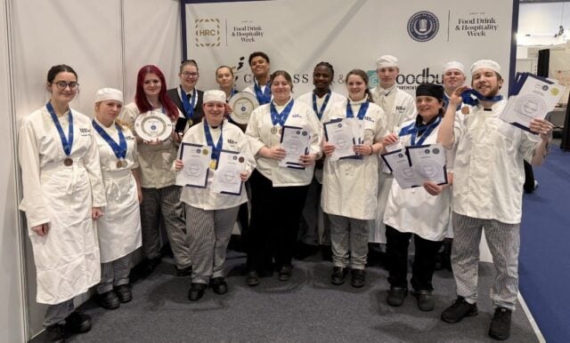 A group of smiling chefs in white uniforms and striped pants proudly display medals and certificates against a backdrop with event logos.