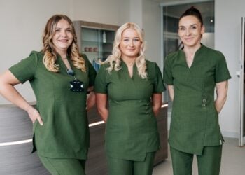 Three women in matching green uniforms stand smiling in an office setting, conveying professionalism and teamwork.