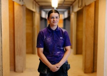 A person wearing a purple polo with "KWL" stands confidently in a corridor. The setting suggests industrial work, with a relaxed, professional atmosphere.