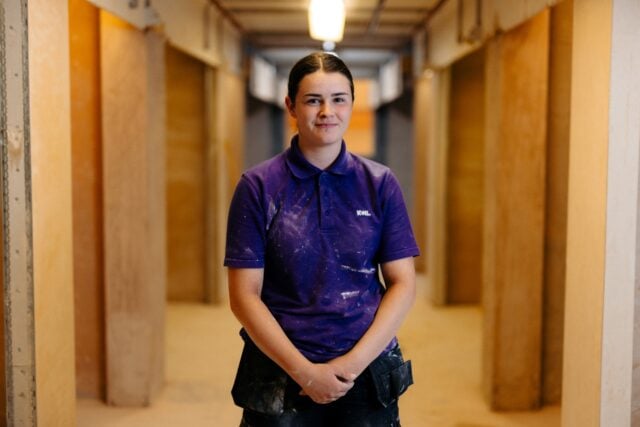 A person wearing a purple polo with "KWL" stands confidently in a corridor. The setting suggests industrial work, with a relaxed, professional atmosphere.