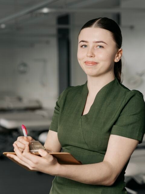 A woman in a green uniform holds a clipboard and pen, smiling softly in a bright, professional setting. She exudes a calm, approachable demeanor.