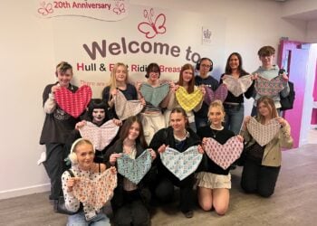 A group of smiling young people stand indoors holding colourful, heart-shaped fabric pieces. Behind them is a wall with a "Welcome to Hull & East Riding Breast Friends" sign.