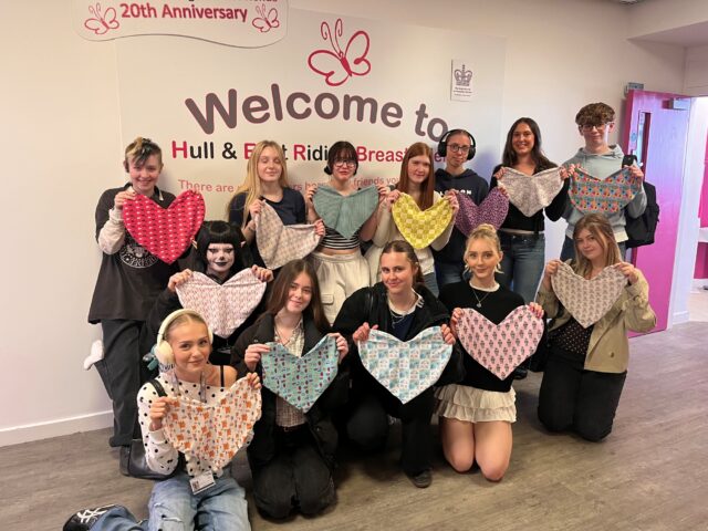 A group of smiling young people stand indoors holding colourful, heart-shaped fabric pieces. Behind them is a wall with a "Welcome to Hull & East Riding Breast Friends" sign.