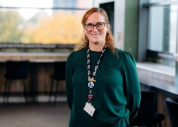 Woman with glasses, wearing a green shirt and lanyard, stands in a modern office with large windows. The atmosphere is professional and warm.