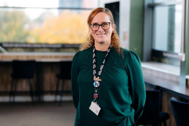 Woman with glasses, wearing a green shirt and lanyard, stands in a modern office with large windows. The atmosphere is professional and warm.