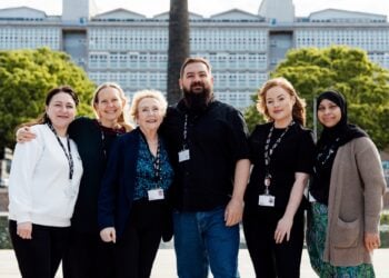 A diverse group of six smiling people stand arm in arm, wearing ID badges. They are outdoors with a large building and green trees in the background, conveying a sense of unity and camaraderie.