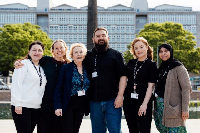 A diverse group of six smiling people stand arm in arm, wearing ID badges. They are outdoors with a large building and green trees in the background, conveying a sense of unity and camaraderie.