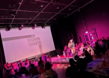 Fashion show with a vibrant pink-lit runway, audience seated at tables. A model in bold attire walks confidently. "The Fashion Show" text on a screen.