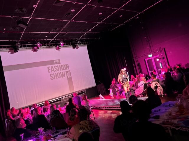 Fashion show with a vibrant pink-lit runway, audience seated at tables. A model in bold attire walks confidently. "The Fashion Show" text on a screen.