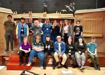 A diverse group of 17 people smiles in a theater set with cardboard houses, creating a warm, collaborative atmosphere. Some are seated on the red steps, others stand behind.