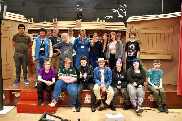 A diverse group of 17 people smiles in a theater set with cardboard houses, creating a warm, collaborative atmosphere. Some are seated on the red steps, others stand behind.