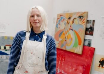 Young artist wearing a blue-striped shirt and paint-splattered overalls stands in a studio, surrounded by colourful abstract paintings. Calm and creative vibe.