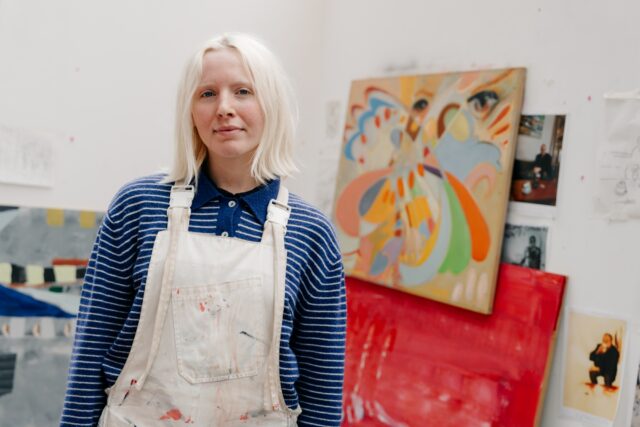 Young artist wearing a blue-striped shirt and paint-splattered overalls stands in a studio, surrounded by colourful abstract paintings. Calm and creative vibe.