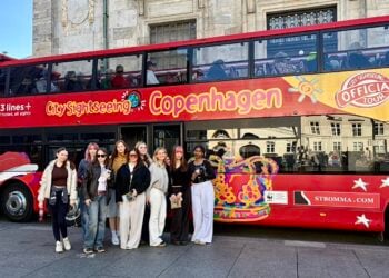 A group of nine people stand smiling in front of a bright red double-decker bus with "Copenhagen City Sightseeing" written on its side. The scene conveys a cheerful, touristy atmosphere.