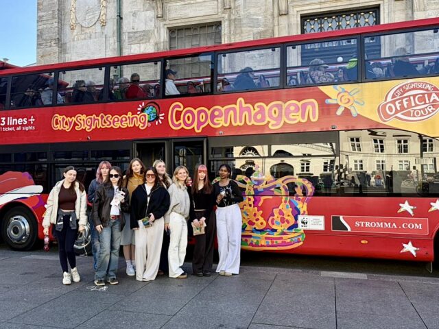A group of nine people stand smiling in front of a bright red double-decker bus with "Copenhagen City Sightseeing" written on its side. The scene conveys a cheerful, touristy atmosphere.