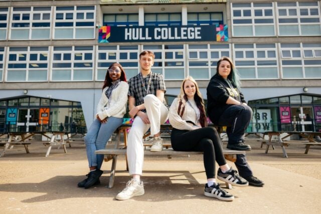 Four students sit and lean on a wooden bench outside Hull College. They appear relaxed and confident, with the college's modern building in the background.