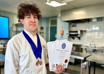 Young chef with curly hair wearing a white uniform and blue medal, holds several certificates, smiling proudly in a stainless steel kitchen.
