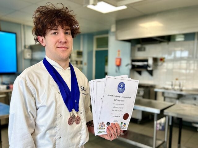 Young chef with curly hair wearing a white uniform and blue medal, holds several certificates, smiling proudly in a stainless steel kitchen.