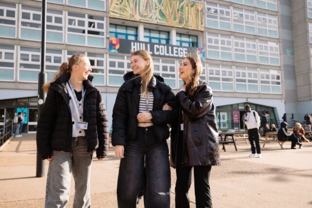 Three smiling students stand arm in arm outside Hull College, wearing winter coats and lanyards, with the modern campus building behind them.