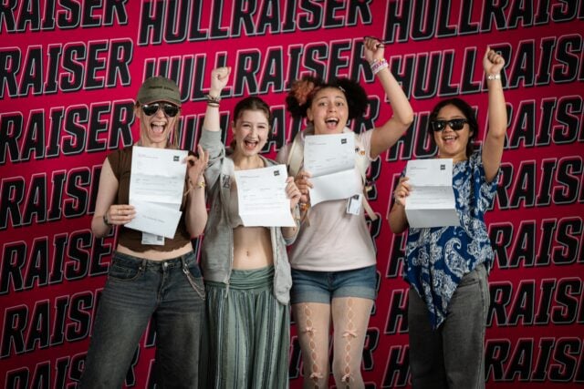Four students hold certificates and raise their fists joyfully against a bold “Hullraisers” backdrop.