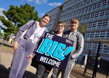 Three smiling young adults pose outside a modern building, holding a colourful “The Big Welcome” sign in bright sunshine.