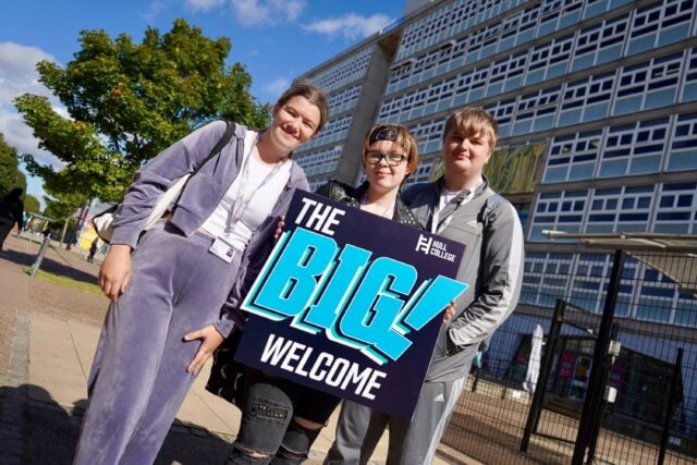 Three smiling young adults pose outside a modern building, holding a colourful “The Big Welcome” sign in bright sunshine.