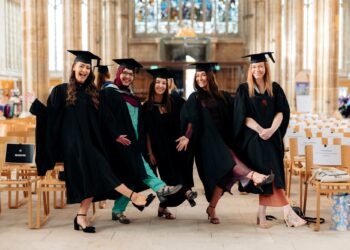 Five joyful graduates in black caps and gowns pose playfully inside a grand cathedral, smiling and kicking up their feet after commencement.
