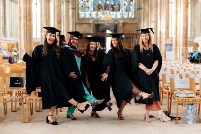 Five joyful graduates in black caps and gowns pose playfully inside a grand cathedral, smiling and kicking up their feet after commencement.