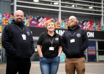 Three smiling staff pose outside a colourful building, wearing black branded clothing and ID badges; friendly, welcoming tone.