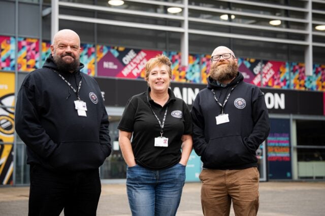 Three smiling staff pose outside a colourful building, wearing black branded clothing and ID badges; friendly, welcoming tone.