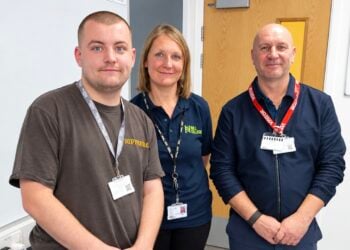 Three people pose together indoors, wearing ID badges and branded work clothing, conveying a friendly, professional atmosphere.