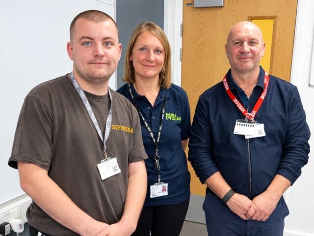 Three people pose together indoors, wearing ID badges and branded work clothing, conveying a friendly, professional atmosphere.