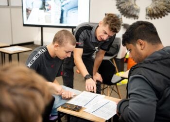 Three men collaborate over printed worksheets in a classroom; an instructor points to a page as two students study it, focused and engaged.