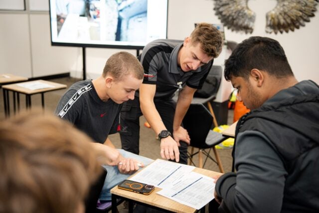 Three men collaborate over printed worksheets in a classroom; an instructor points to a page as two students study it, focused and engaged.