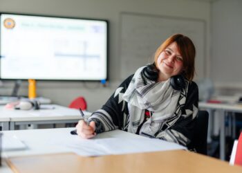 A smiling woman with headphones around her neck writes at a classroom desk, with a blurred presentation screen behind her; the mood is warm and relaxed.