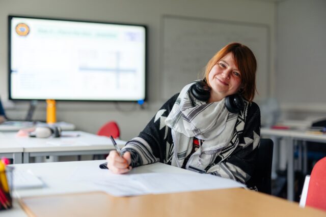 A smiling woman with headphones around her neck writes at a classroom desk, with a blurred presentation screen behind her; the mood is warm and relaxed.