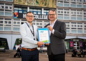 Two smiling men stand outside a college building, jointly holding a blue-framed certificate for the camera; the scene feels celebratory and professional.