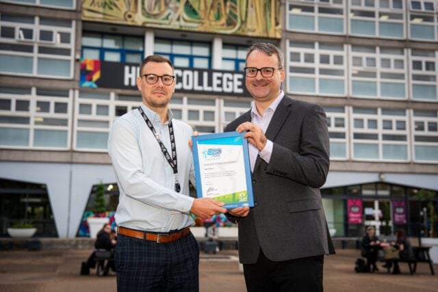 Two smiling men stand outside a college building, jointly holding a blue-framed certificate for the camera; the scene feels celebratory and professional.