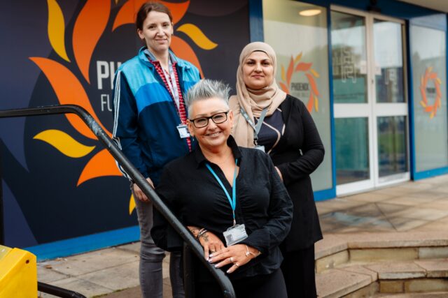 Three women pose outside a building with a flame mural and glass doors; one smiles in front as two stand behind, conveying warmth and confidence.