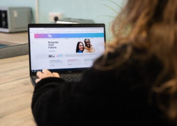 A person in a black sweater browses a career website on a laptop at a wooden desk, viewing a “Reignite Your Future” page with a smiling couple.