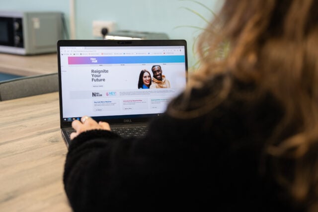 A person in a black sweater browses a career website on a laptop at a wooden desk, viewing a “Reignite Your Future” page with a smiling couple.